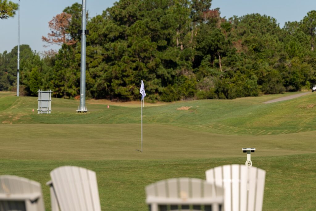 White Adirondack chairs beside a green on The Piper, with a pin flag and light poles beyond