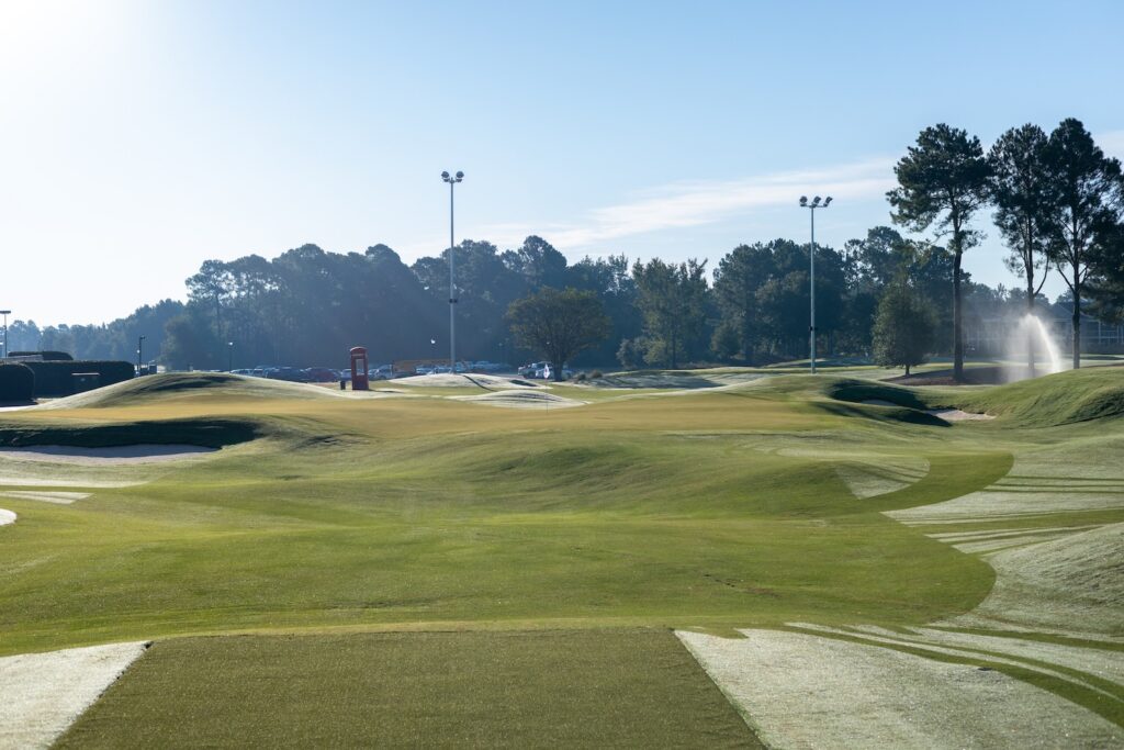 The first tee of The Piper on a dewy morning, with mounded greens, a bunker and the red telephone box in the distance