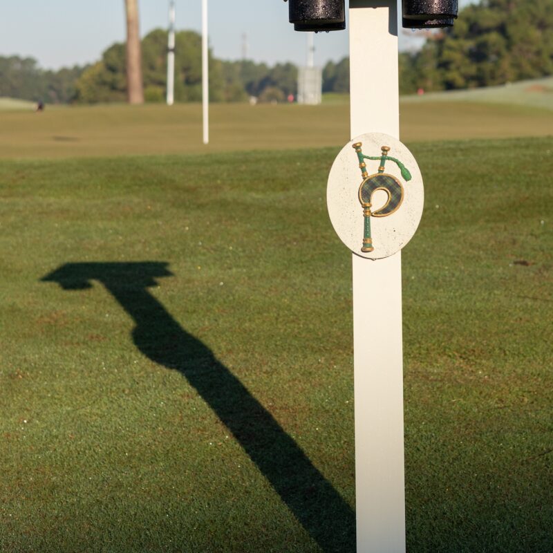 A white tee marker post with a tartan bagpipe badge, casting a long shadow across the tee