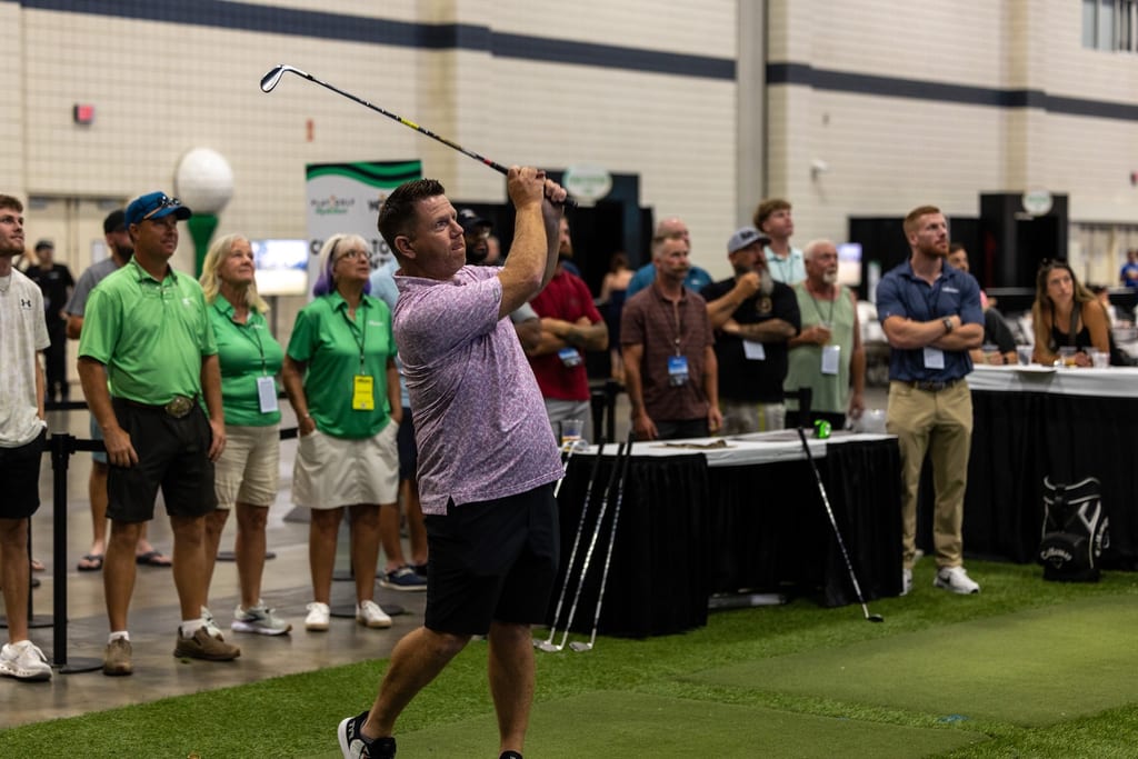 Players hitting shots off turf on the convention floor while a crowd watches from the bar