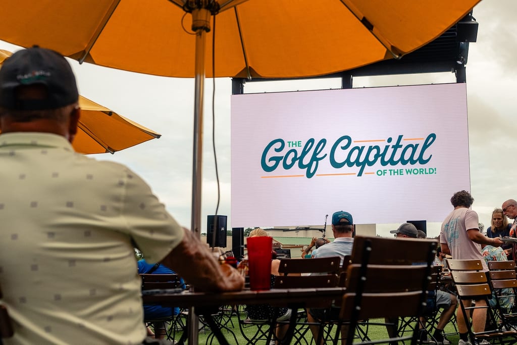 Players and guests seated at outdoor tables in front of a large video screen