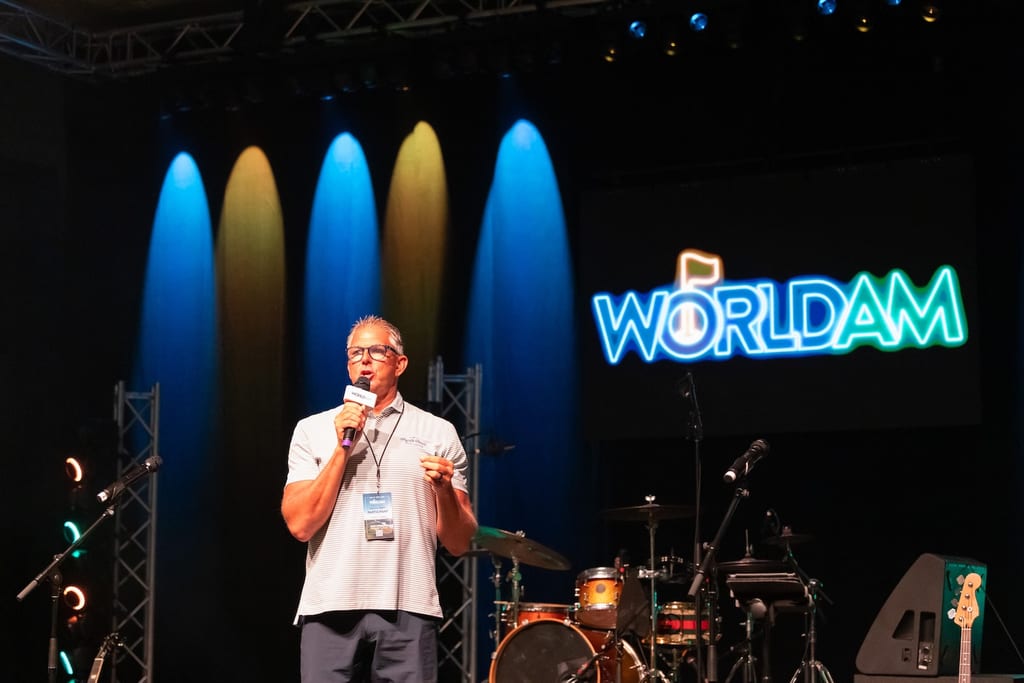 A host with a World Am microphone on the lit ballroom stage in front of the band setup